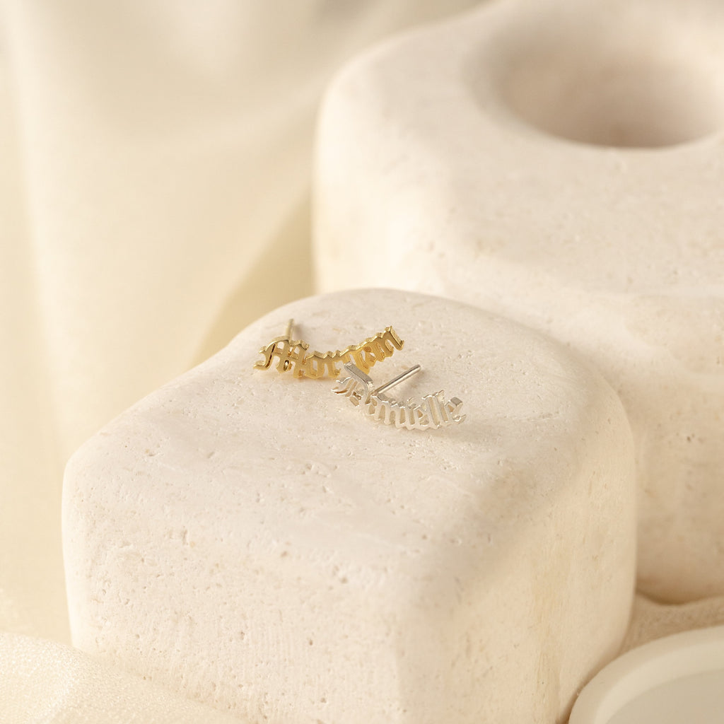 Old English Name Earrings rest on cream-colored textured stone blocks under soft, neutral lighting, making them a perfect custom gift.