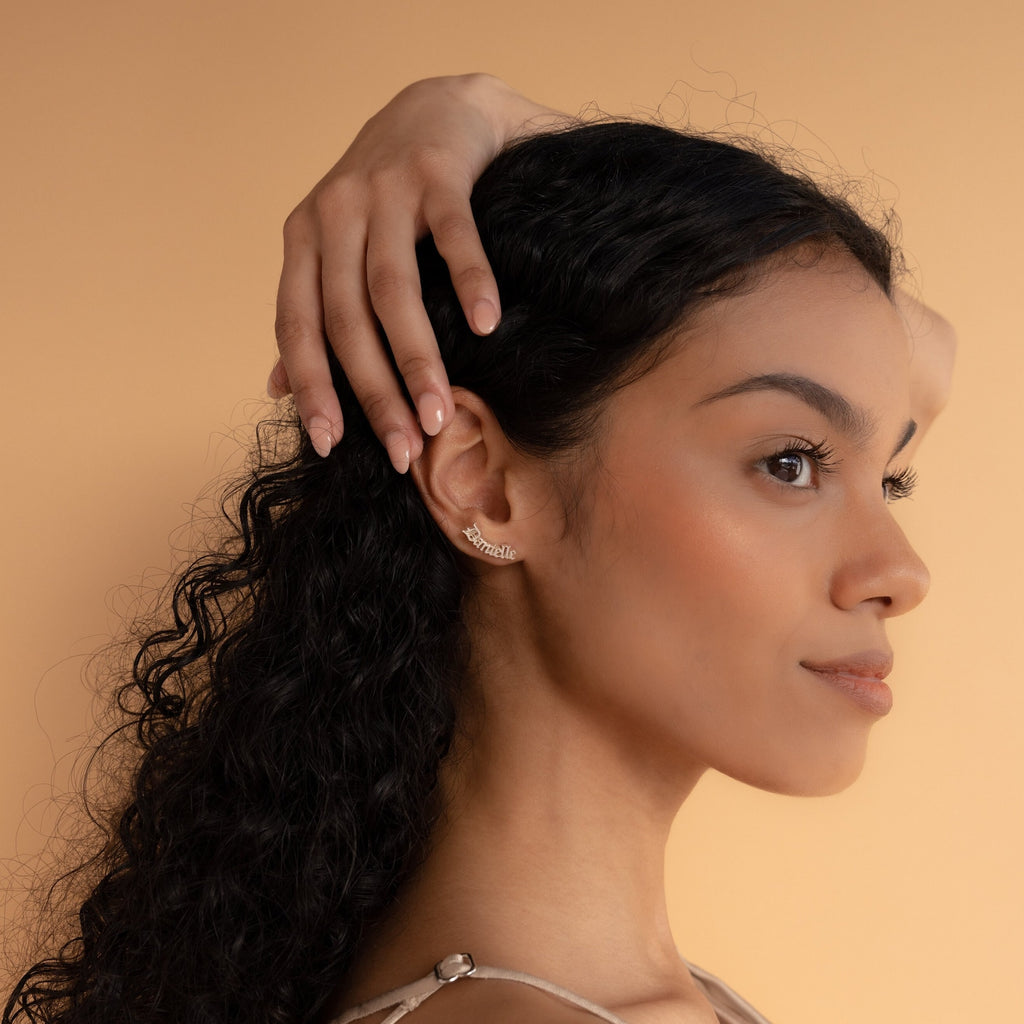 Woman with long curly hair poses sideways, hand on head, wearing elegant Old English Name Earrings—a perfect custom gift—against a beige background.