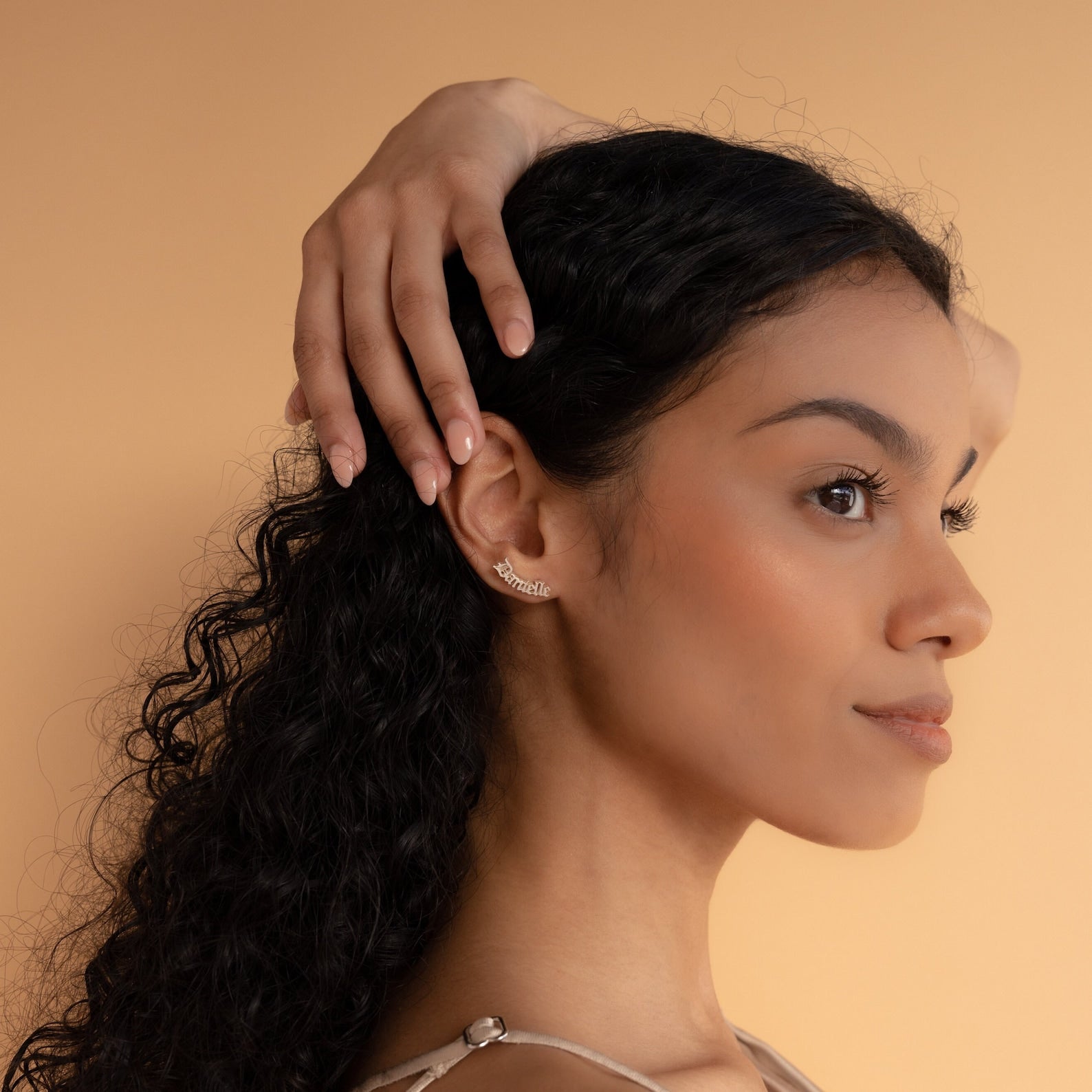 Woman with long curly hair poses sideways, hand on head, wearing elegant Old English Name Earrings—a perfect custom gift—against a beige background.