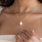 A woman gently touches her chest while wearing the Ariel Pearl Necklace, a gold chain with a single white pearl pendant—an elegant choice for bridesmaid jewelry.