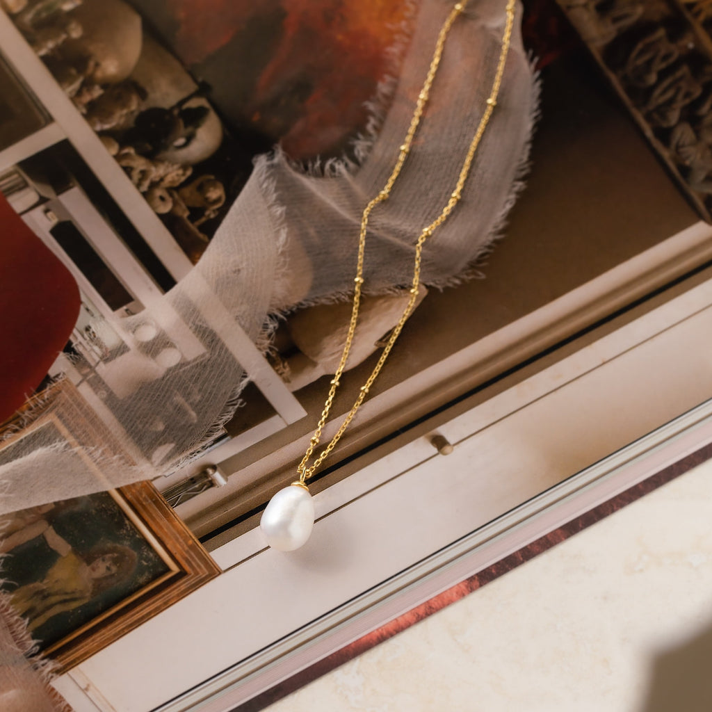 The Ariel Pearl Necklace features a gold chain with a delicate pearl pendant, displayed on vintage photos and fabric—perfect as bridal jewelry or a thoughtful bridesmaid gift.