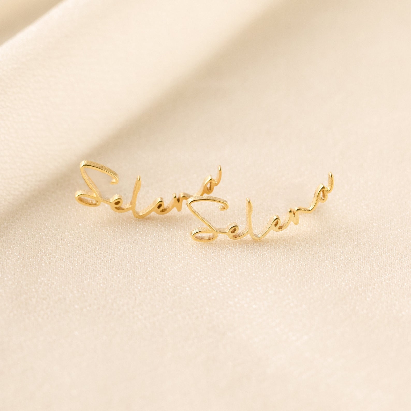 Two Summer Name Earrings rest on soft, cream-colored fabric. Each earring features elegant cursive script, spelling Selena—a stunning example of personalized jewelry.