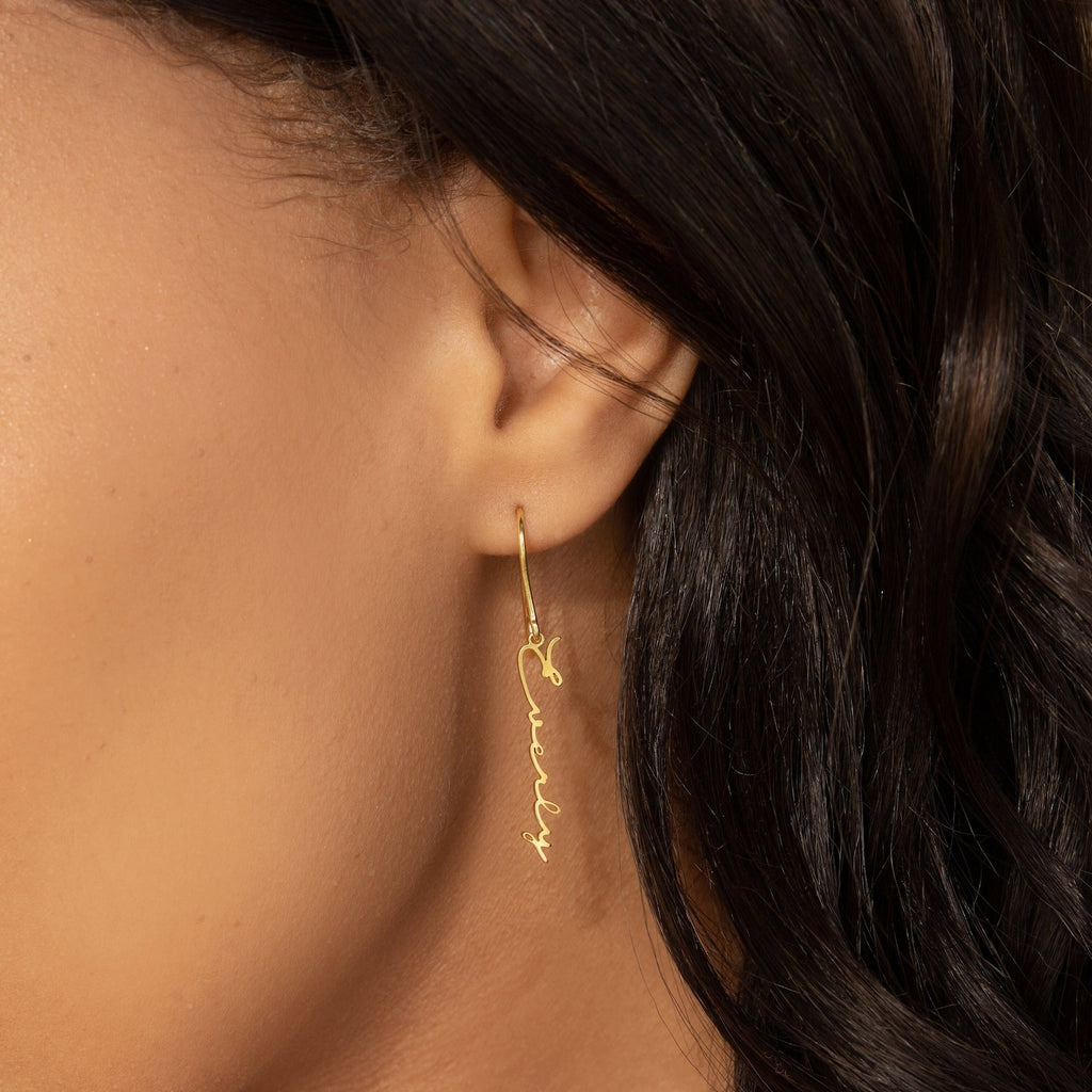A gold Minimal Dangling Name Earring spelling "curly" hangs from the ear of a woman with wavy dark hair—an elegant bridesmaid gift.