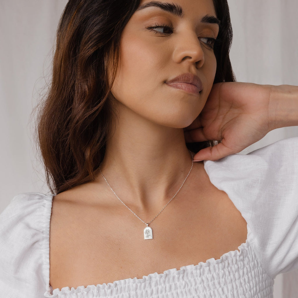 Woman in a white top wears the Birth Flower Arch Necklace with a silver pendant, looking to the side with one hand behind her neck.