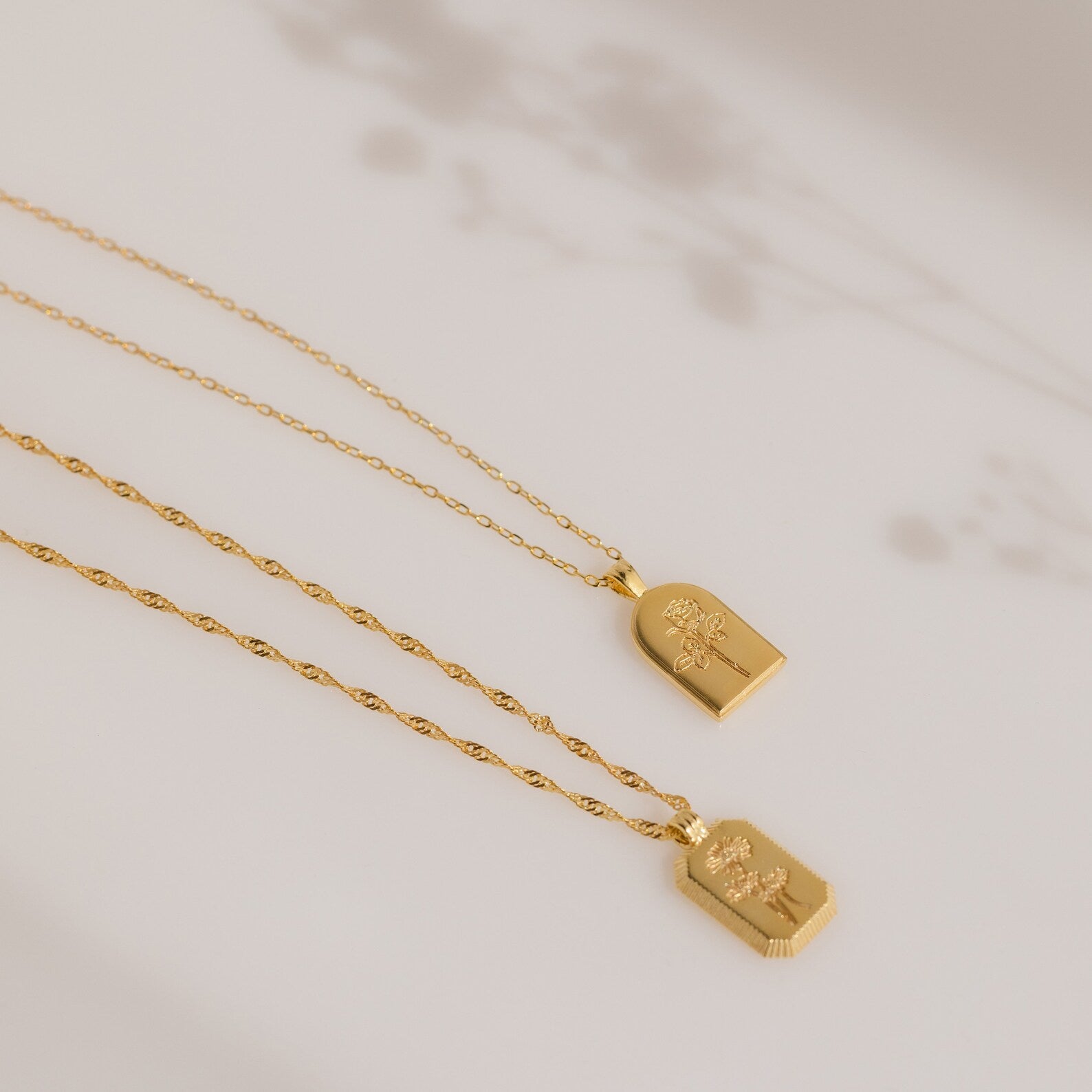 Two Birth Flower Arch Necklaces with delicate chains rest on a neutral background with soft floral shadows—perfect personalized jewelry or a cherished birth flower necklace.