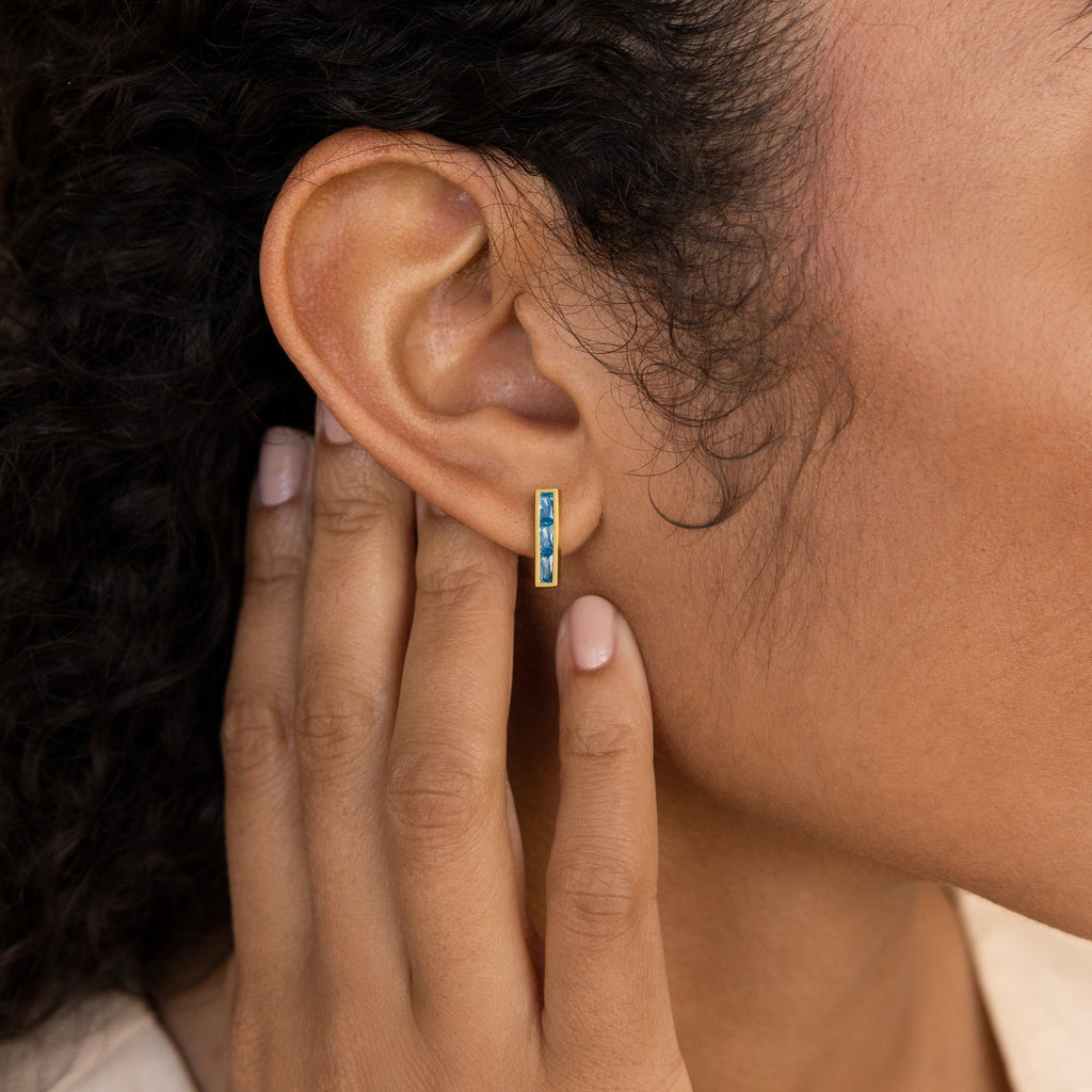 Close-up of a person touching their ear, wearing Baguette Birthstone Bar Studs—small gold earrings featuring blue stones.