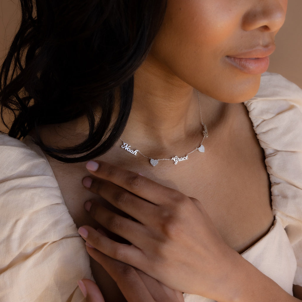 A woman in a cream dress touches the Mellow Name Heart Necklace, a dainty chain featuring personalized “Noah” and “Grace” name charms with hearts.
