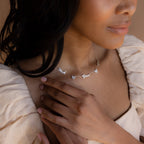 A woman in a cream dress touches the Mellow Name Heart Necklace, a dainty chain featuring personalized “Noah” and “Grace” name charms with hearts.