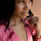 A woman in a pink top smiles softly, looking to the side while wearing a minimalist Old English Letter Name Necklace.