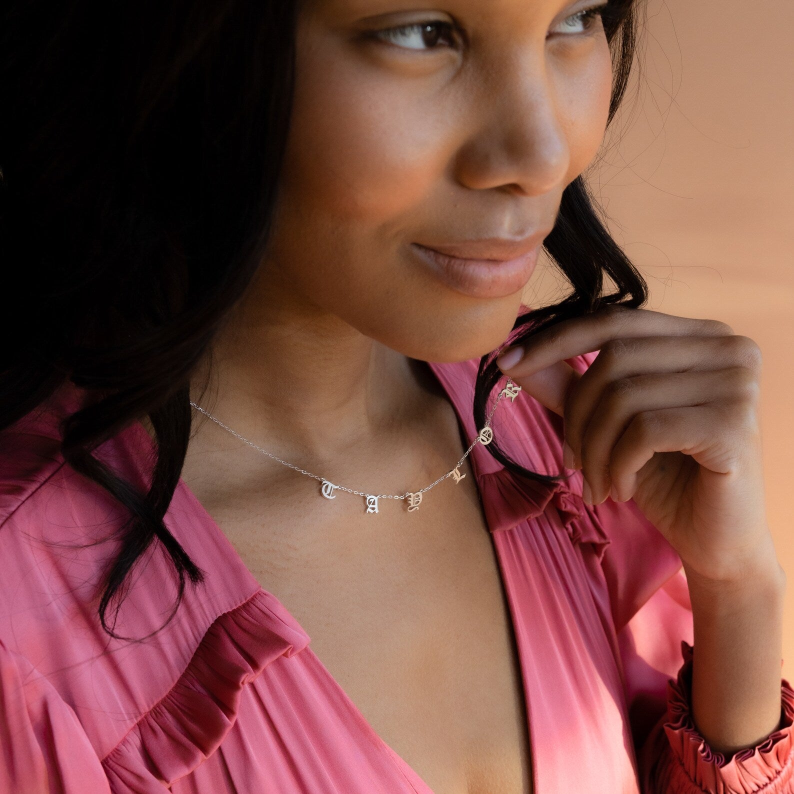 A woman in a pink top smiles softly, looking to the side while wearing a minimalist Old English Letter Name Necklace.