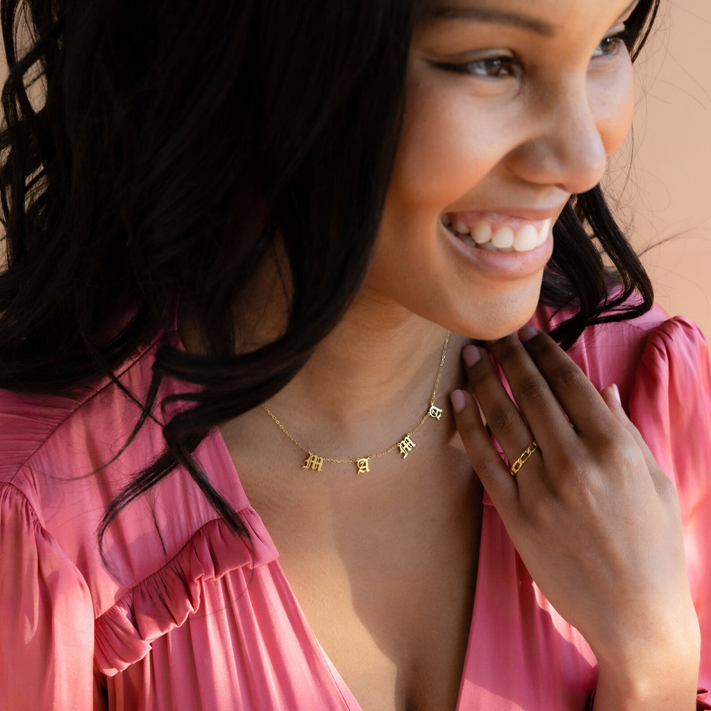 A smiling woman in a pink dress wears the Old English Letter Name Necklace paired with a matching minimalist ring.