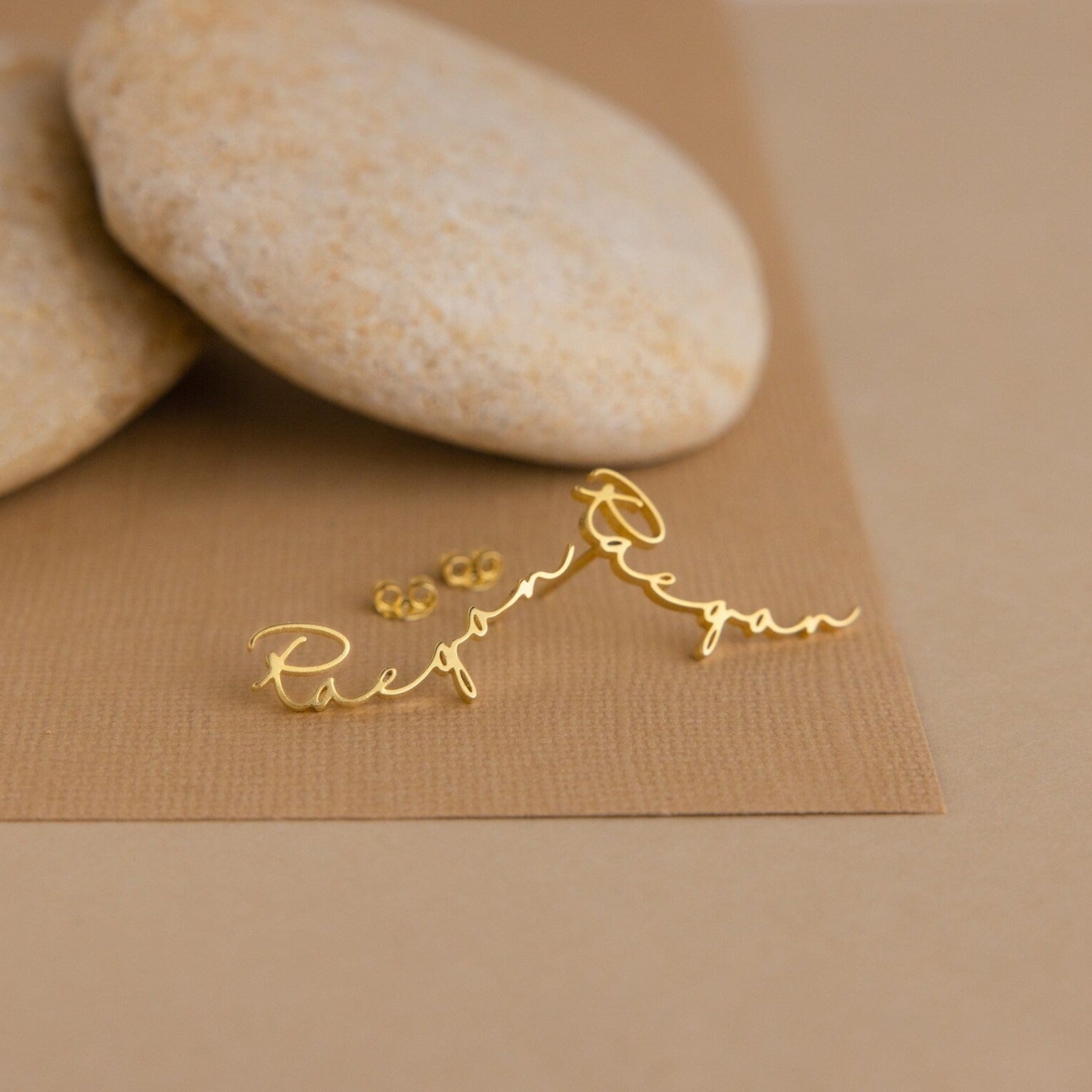 Gold personalized Hallie Name Earrings in cursive, shown on a brown background with smooth stones—ideal as a custom gift or unique name stud earrings.