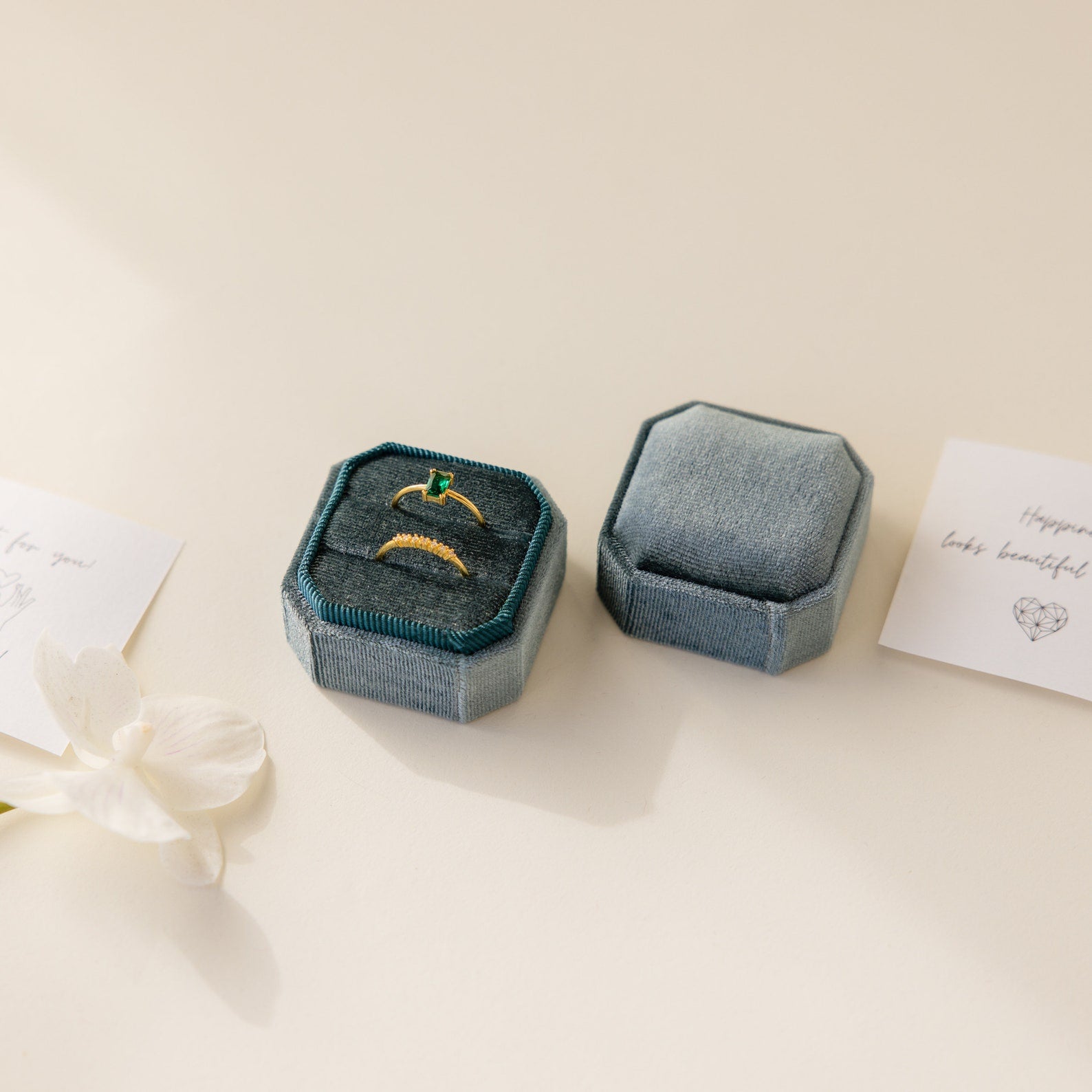 A 2 Slot Velvet Ring Box holding a gold and green ring beside its lid, a flower, and handwritten notes.