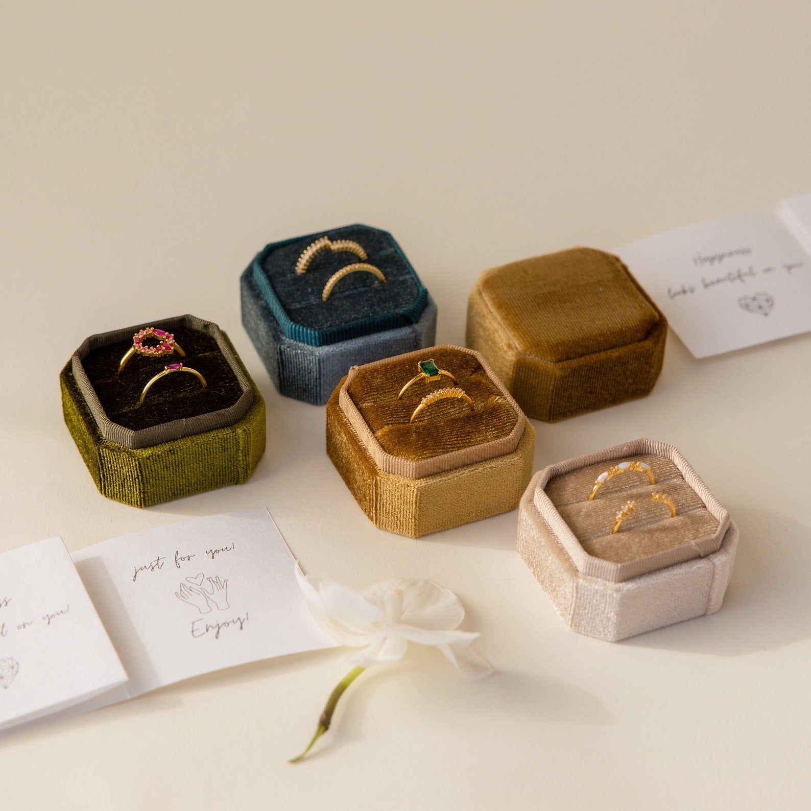 Five elegant 2 Slot Velvet Ring Boxes are displayed with rings, cards, and a white flower on a beige surface.