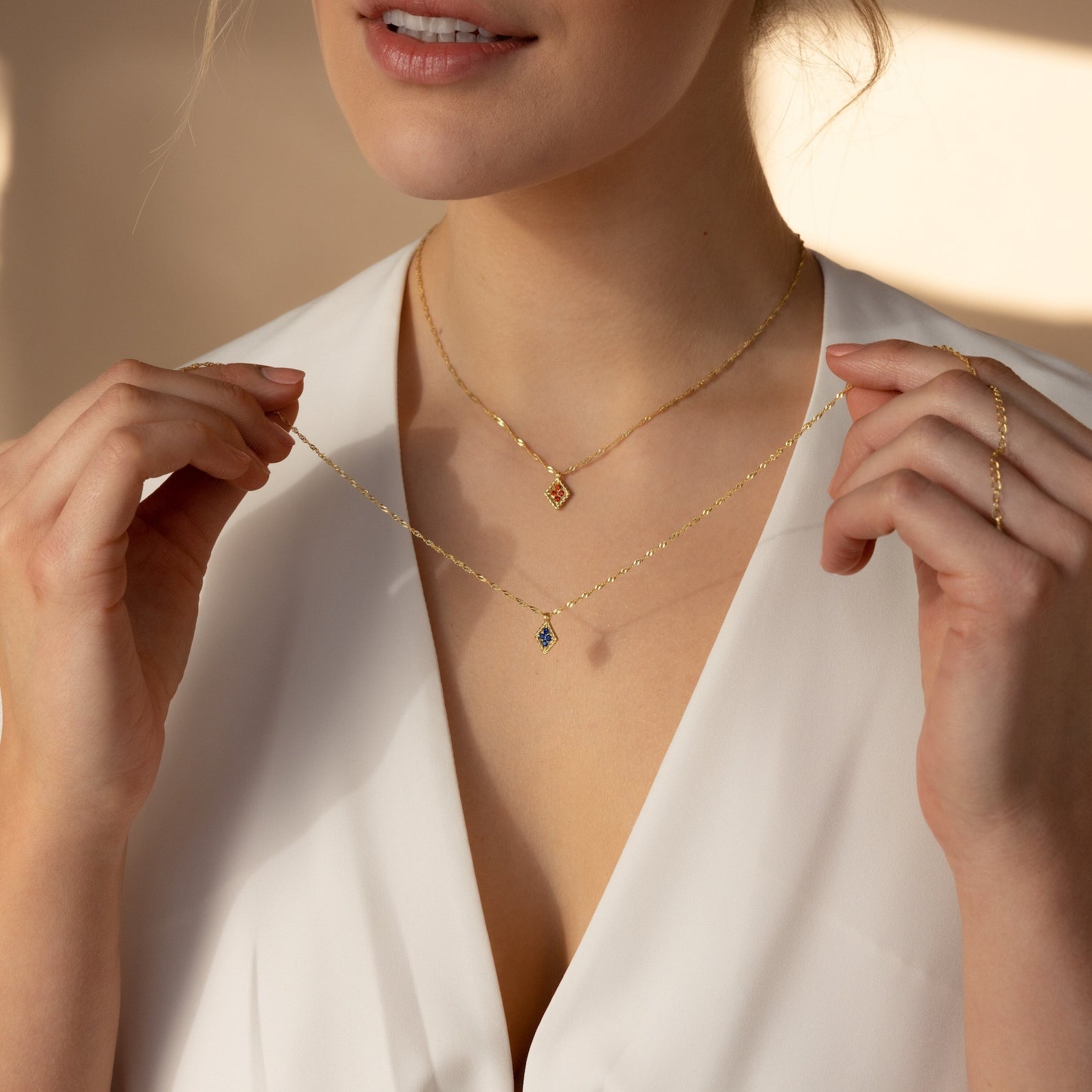 A woman in a white blouse showcases two gold necklaces, including the Birthstone Diamond Pendant Necklace—a handmade piece featuring gemstones, small pendants, and subtle personalized jewelry details.