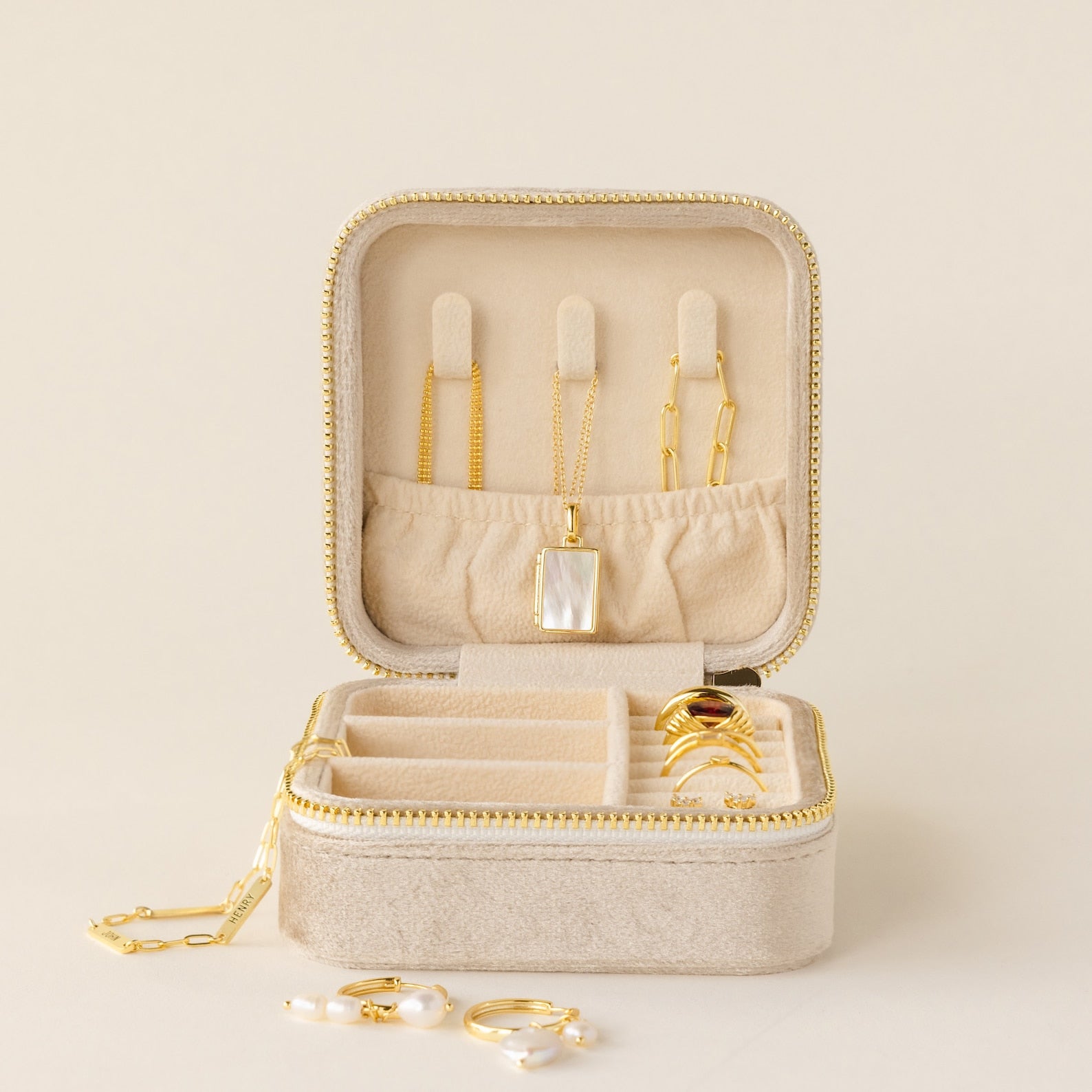 A Square Velvet Jewelry Case in beige velvet displays gold rings, necklaces, a bracelet, and pearl earrings inside and around it.