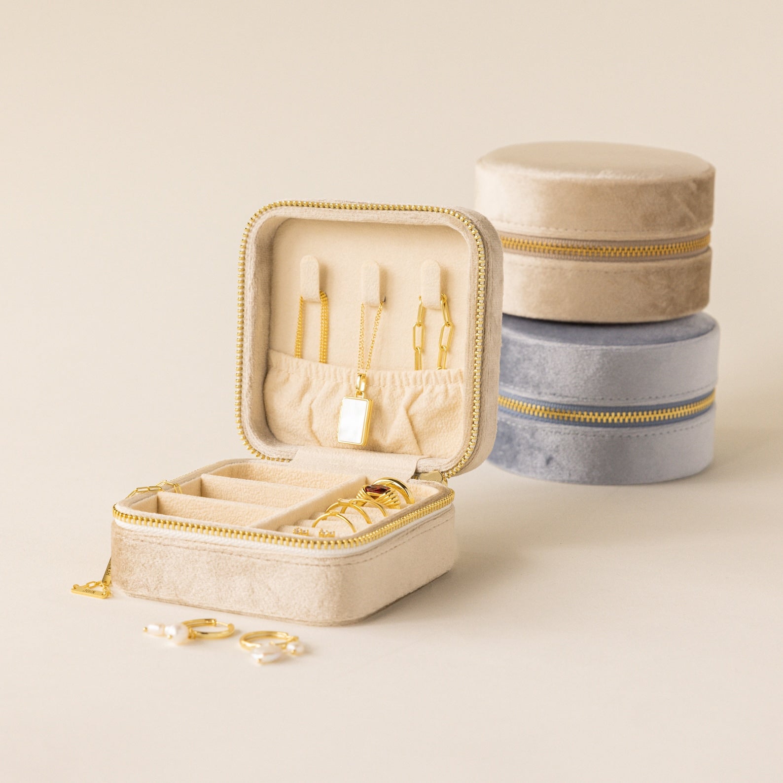 Square Velvet Jewelry Case in beige velvet, shown open with gold jewelry inside, placed next to closed beige and gray cases on a neutral background.