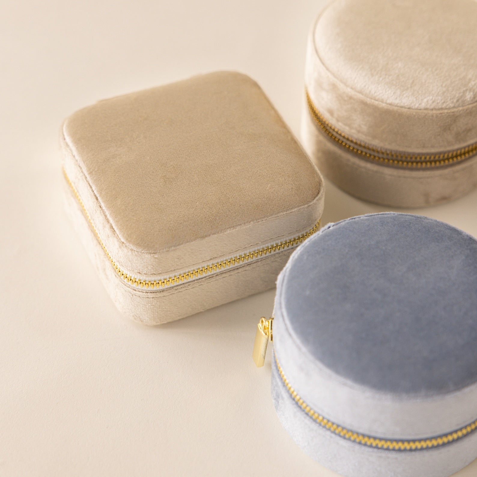Three Square Velvet Jewelry Cases in beige and blue with gold zippers are shown on a light surface—elegant jewelry boxes ideal for travel.