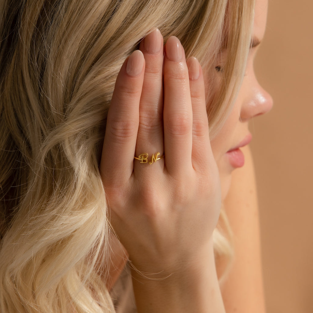 A blonde woman wears the Hallie Duo Initial Ring, featuring gold BN lettering on her finger while covering part of her face—an elegant, personalized jewelry piece.