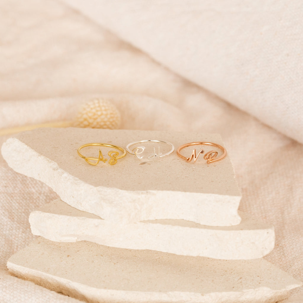 The Hallie Duo Initial Ring, shown in gold, silver, and rose gold on stone slabs against a beige backdrop, makes a perfect personalized jewelry gift for mothers.