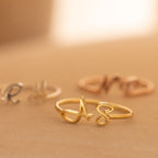 Close-up of Hallie Duo Initial Rings in gold, silver, and rose gold with cursive initials on a soft beige surface—personalized jewelry that makes a thoughtful gift for mothers.