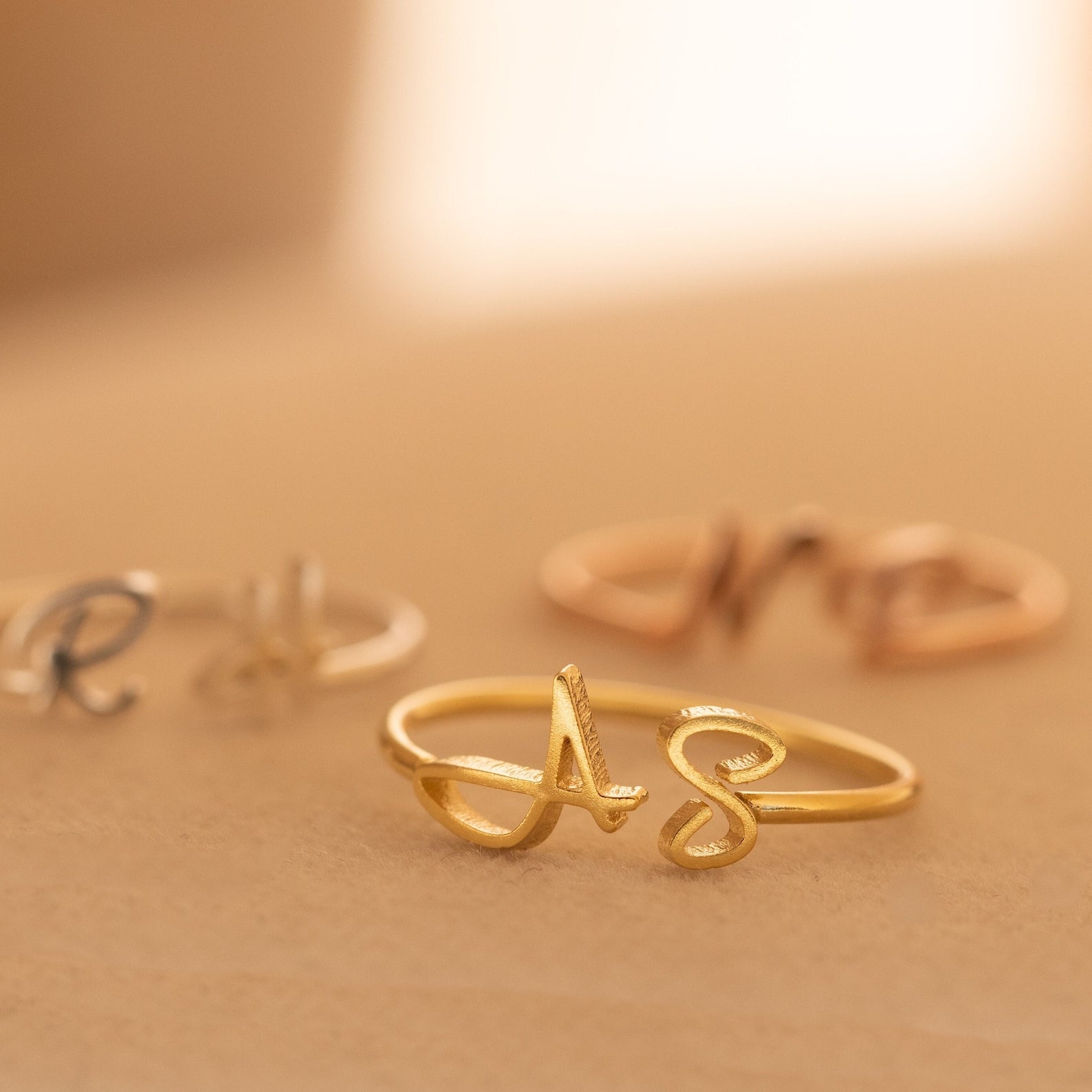 Close-up of Hallie Duo Initial Rings in gold, silver, and rose gold with cursive initials on a soft beige surface—personalized jewelry that makes a thoughtful gift for mothers.