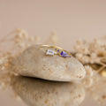 The Space Baguette Birthstone Ring with clear and purple stones rests on a smooth stone, with softly blurred dried flowers in the background.