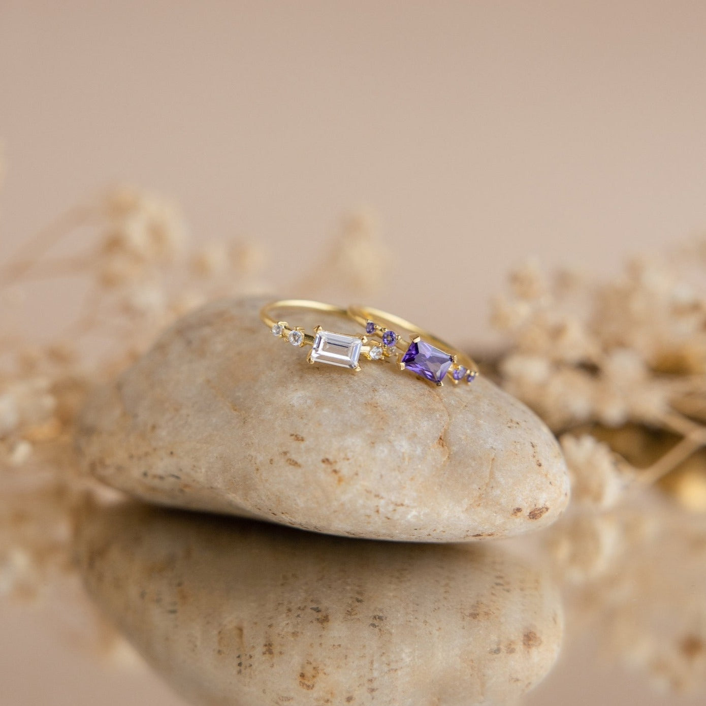 The Space Baguette Birthstone Ring with clear and purple stones rests on a smooth stone, with softly blurred dried flowers in the background.