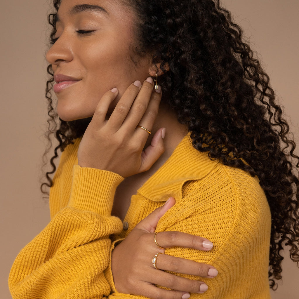 Woman in yellow sweater with curly hair smiles softly, eyes closed, as she gently touches her earrings and the Space Baguette Birthstone Ring on her finger.