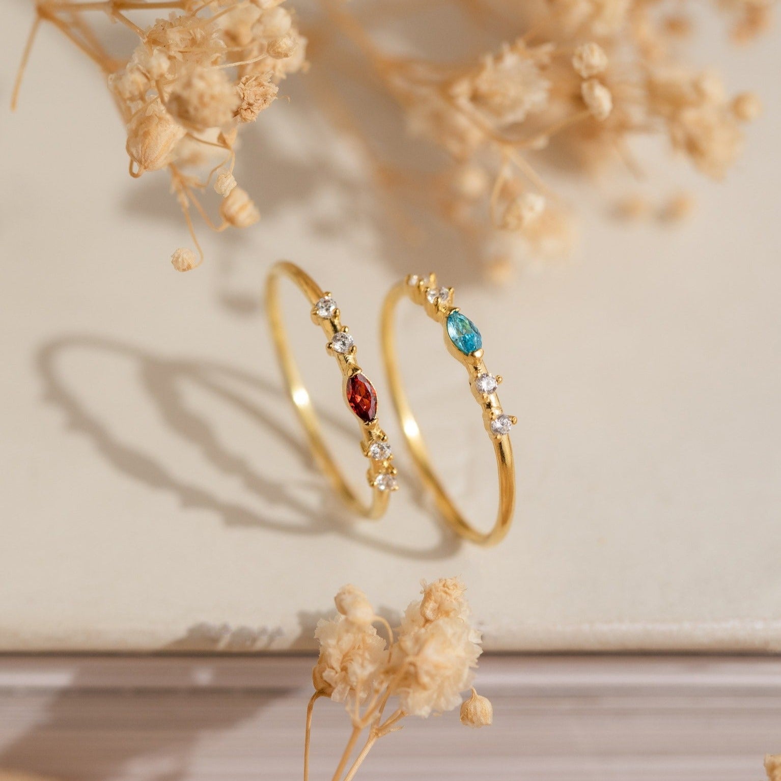 The Space Marquise Birthstone Ring, along with two gold rings accented with small white stones and red and blue gems, glimmers among dried flowers on a neutral surface.