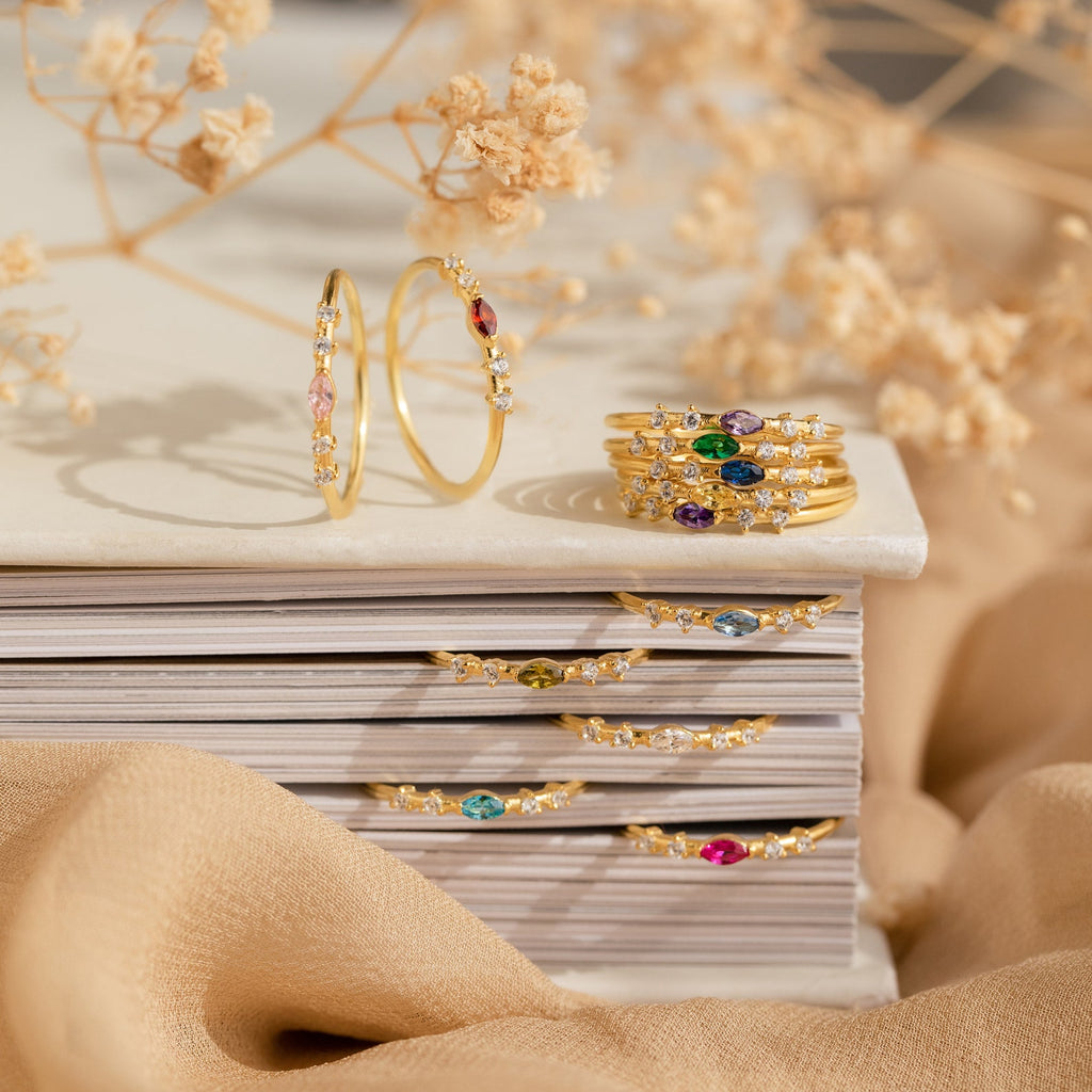 The Space Marquise Birthstone Ring, featuring customizable gemstones and colorful crystals, is displayed on stacked books with dried flowers in the background.