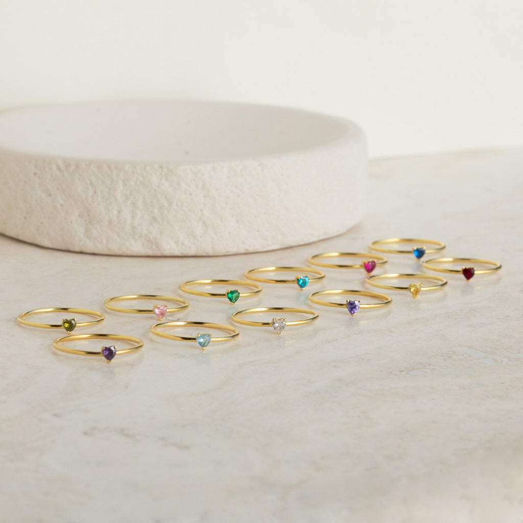 The Tiny Heart Birthstone Ring, a gold stacking ring featuring a heart shape and colorful gemstone, is displayed on light marble near a round slab—an ideal romantic gift.
