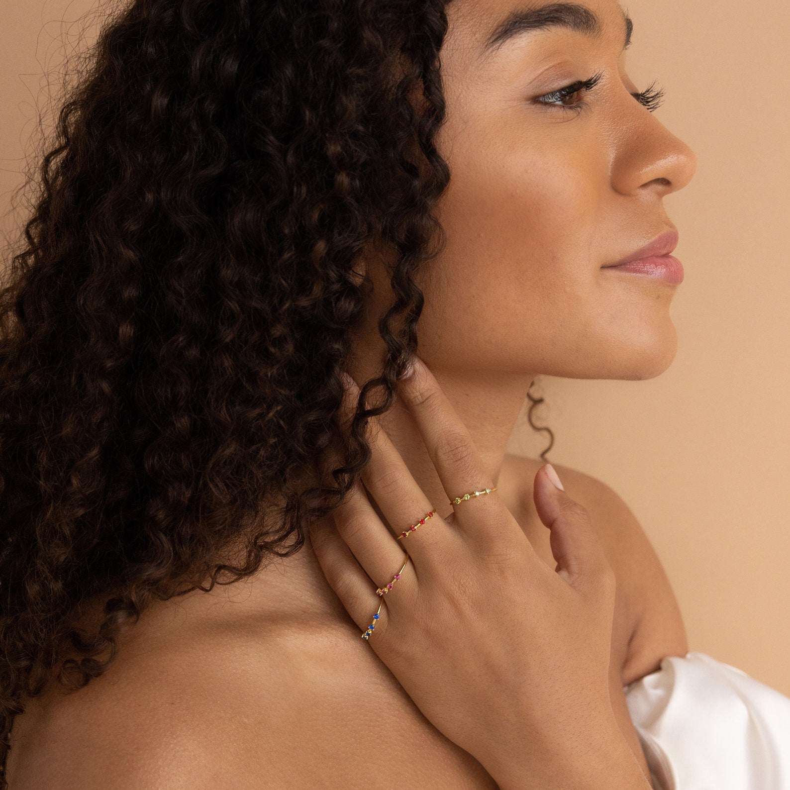 A woman with curly hair poses in profile, wearing the Space Birthstone Ring stacked with beaded rings, paired with a white off-shoulder top against a beige background.