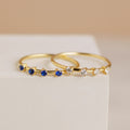 The Space Birthstone Ring, a delicate gold band featuring small blue stones, rests elegantly on a white surface.