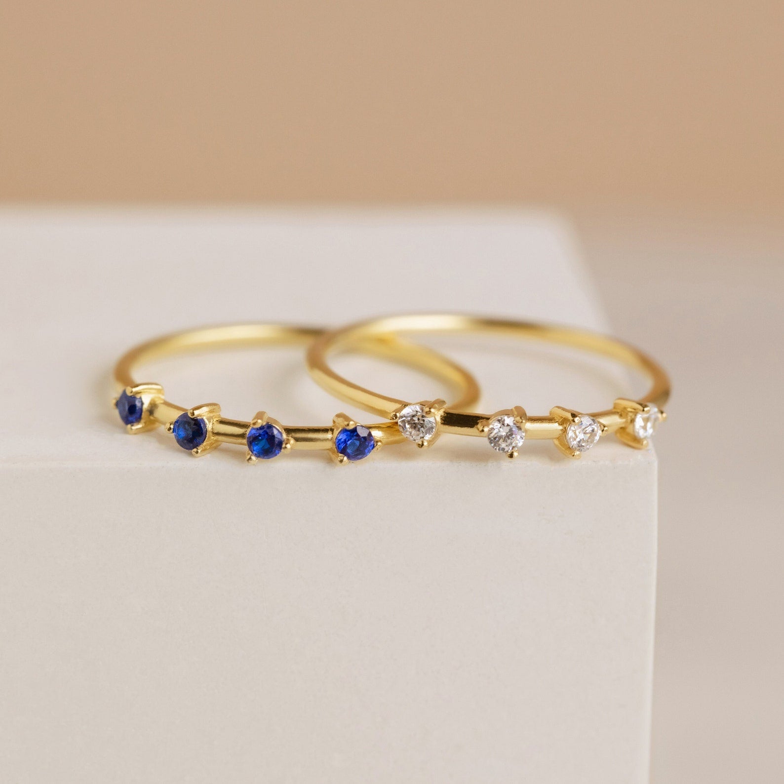 The Space Birthstone Ring, a delicate gold band featuring small blue stones, rests elegantly on a white surface.