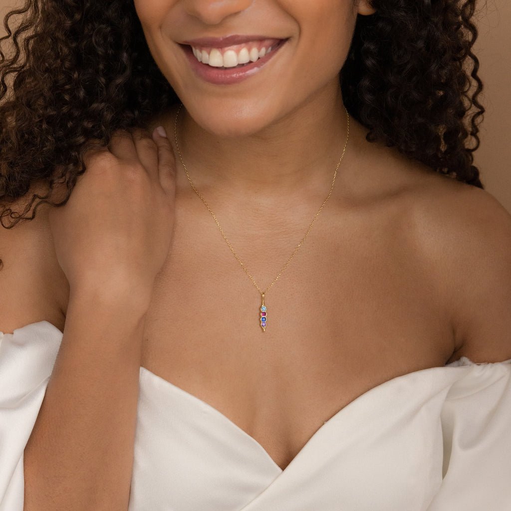 Smiling woman in a white off-shoulder top wearing a gold Vertical Pod Birthstone Necklace with a colorful, personalized pendant.