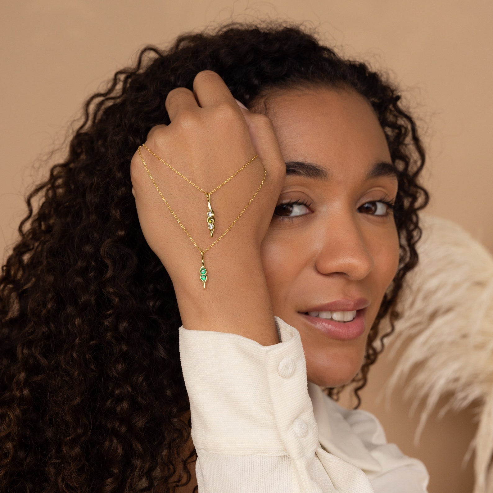 A woman with curly hair raises her hand, displaying a Vertical Pod Birthstone Necklace—custom gold jewelry featuring green and clear gemstone pendants for those who love personalized accessories.