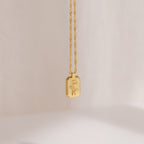 The Ribbed Flower Tag Necklace is a vintage-inspired piece featuring a rectangular gold charm engraved with flowers, elegantly hanging from a twisted chain.