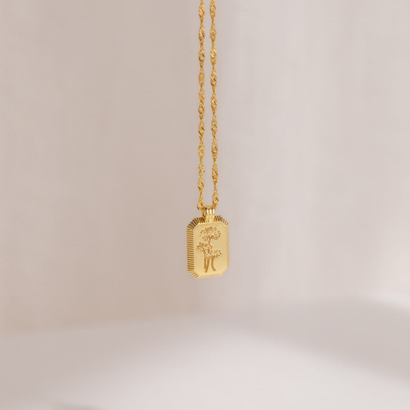 The Ribbed Flower Tag Necklace is a vintage-inspired piece featuring a rectangular gold charm engraved with flowers, elegantly hanging from a twisted chain.