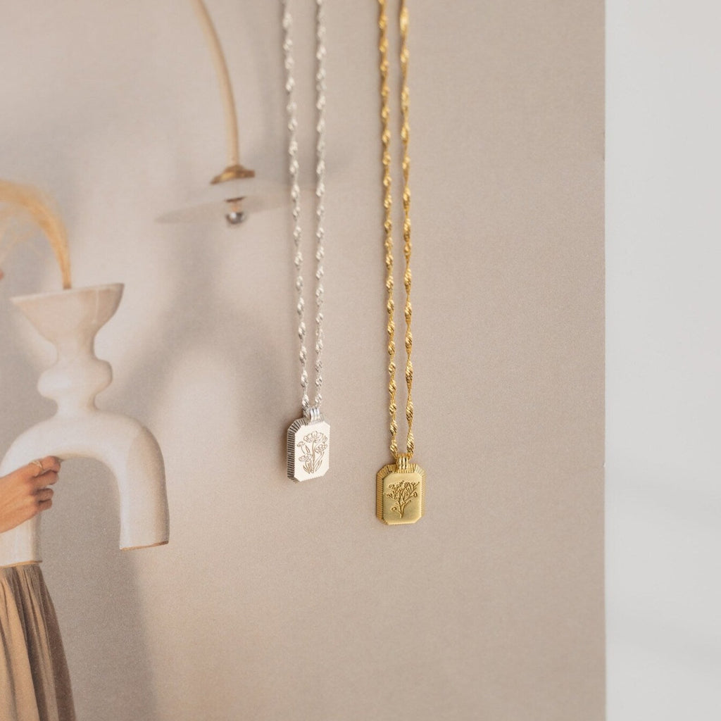 Silver and gold necklaces, including the Ribbed Flower Tag Necklace with engraved pendants, are displayed against a soft, neutral-toned background.
