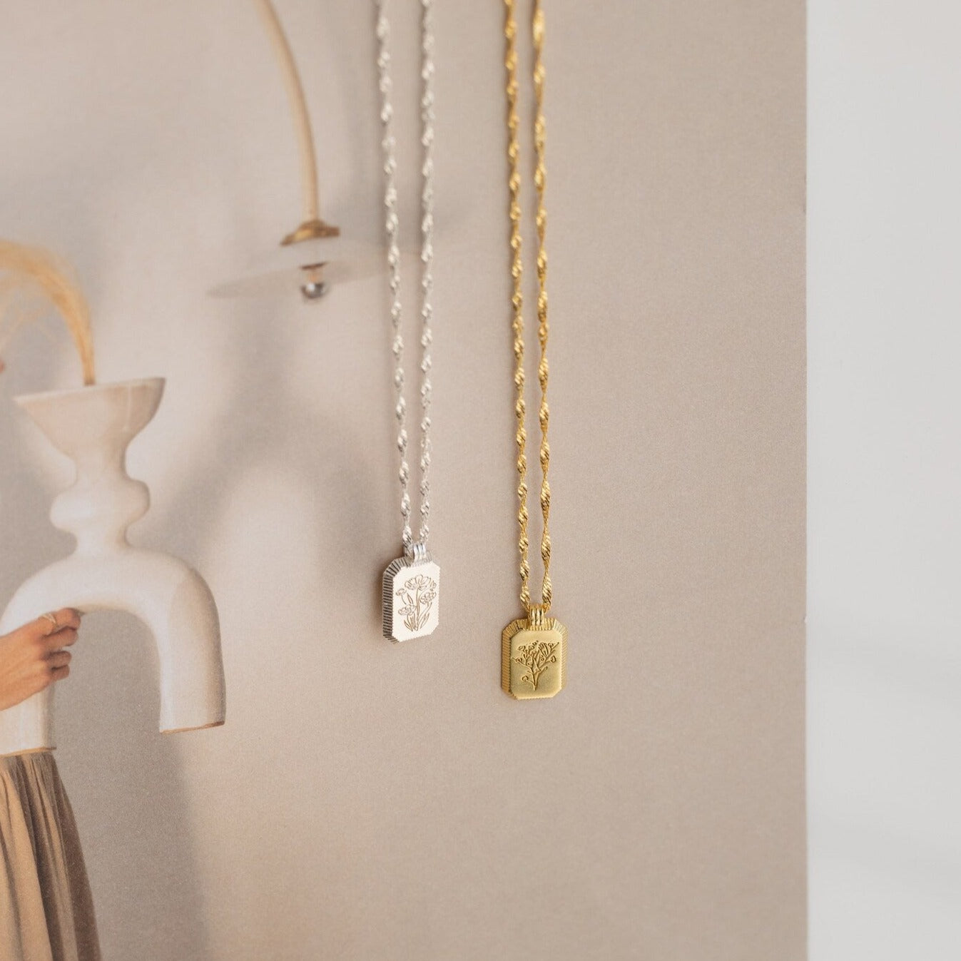 Silver and gold necklaces, including the Ribbed Flower Tag Necklace with engraved pendants, are displayed against a soft, neutral-toned background.