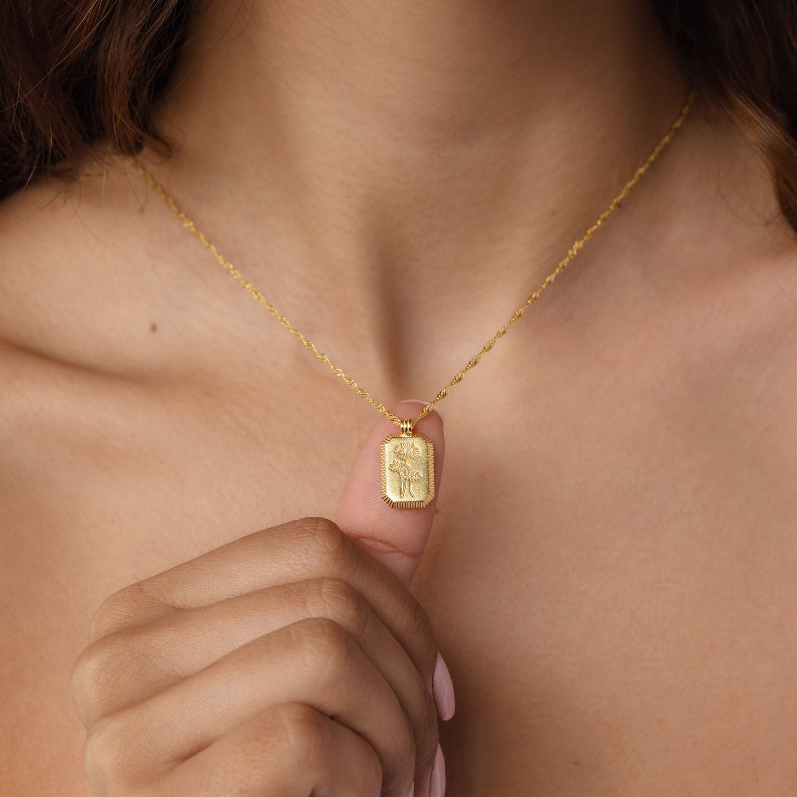 A close-up of a person holding the Ribbed Flower Tag Necklace between their fingers, showcasing its rectangular pendant. This vintage-inspired piece adds an elegant charm to any outfit.