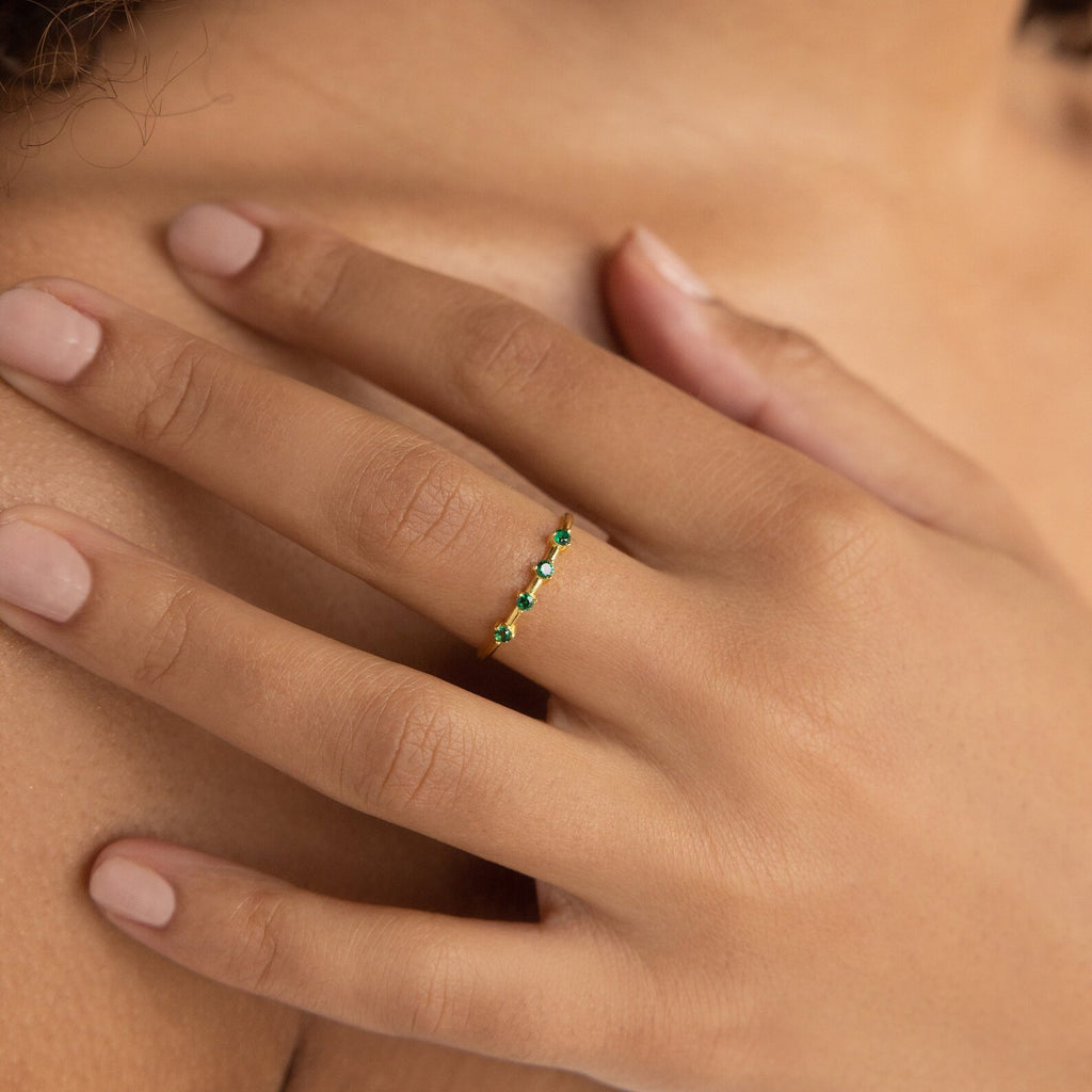 A manicured hand wears the Space Birthstone Ring, a delicate gold band adorned with small green gemstones.