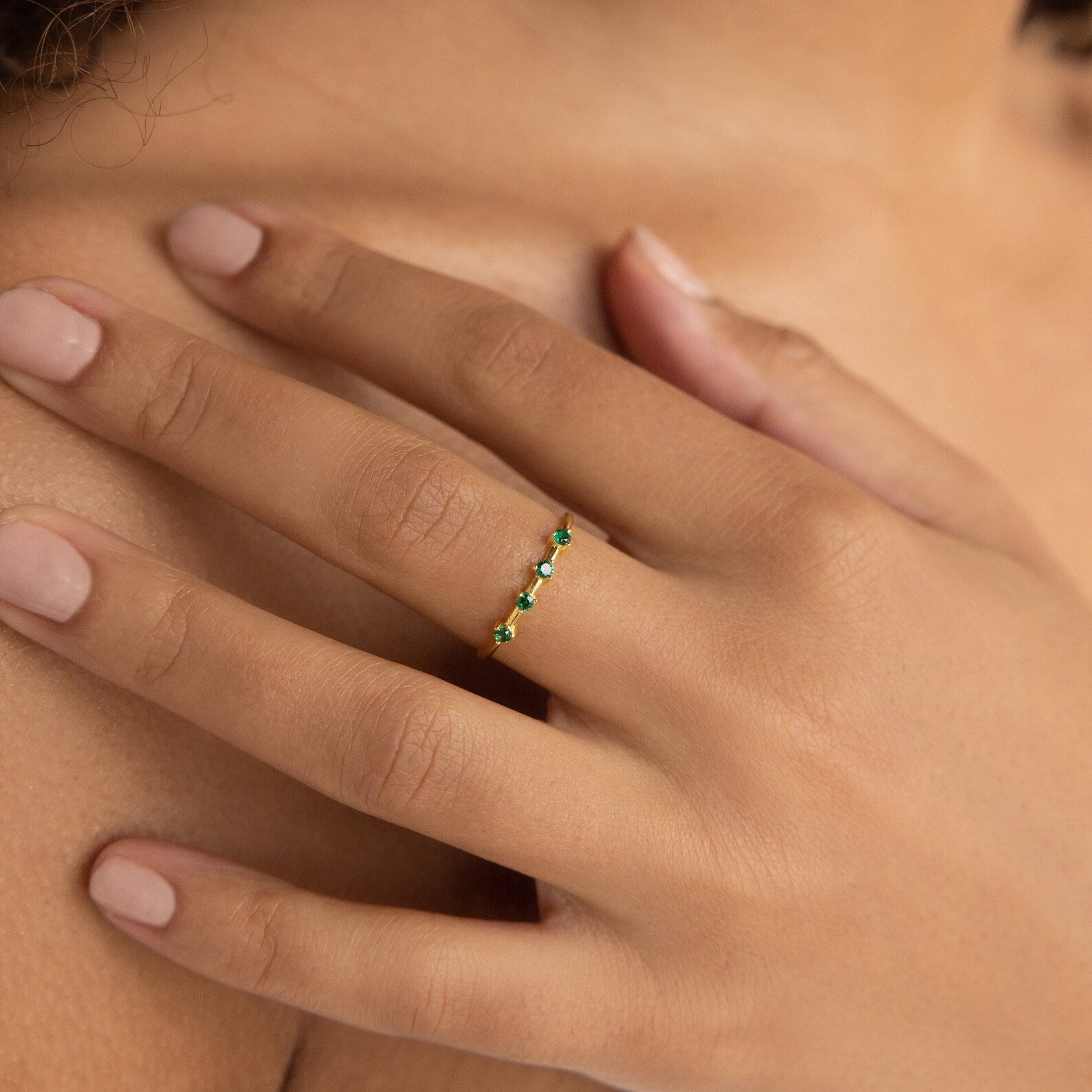 A manicured hand wears the Space Birthstone Ring, a delicate gold band adorned with small green gemstones.