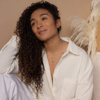 A woman with curly hair in a cream shirt sits against a beige background with pampas grass, wearing unique layered necklaces including the Horizontal Pod Birthstone Necklace.
