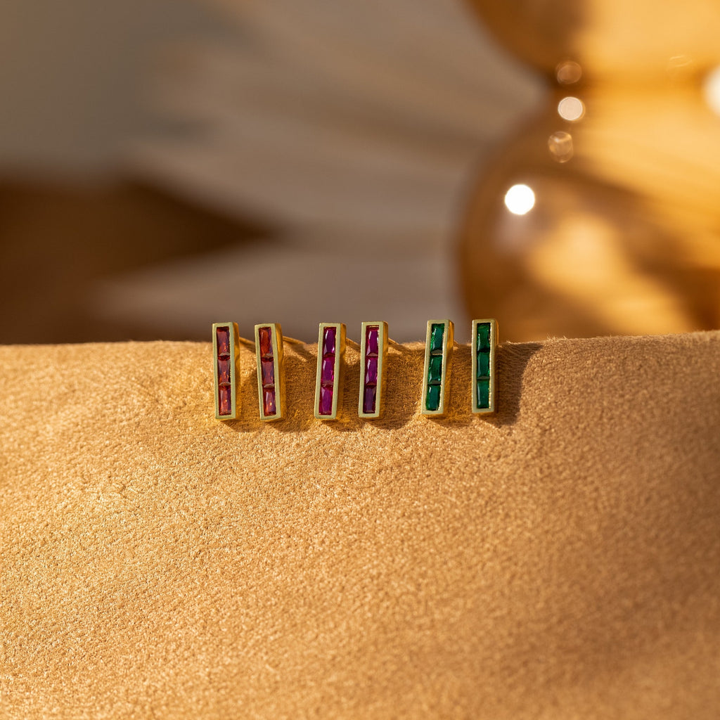 Three pairs of Baguette Birthstone Bar Studs in gold, featuring red, purple, and green stones, rest on a soft tan surface—perfect for adding color to any ear piercing.