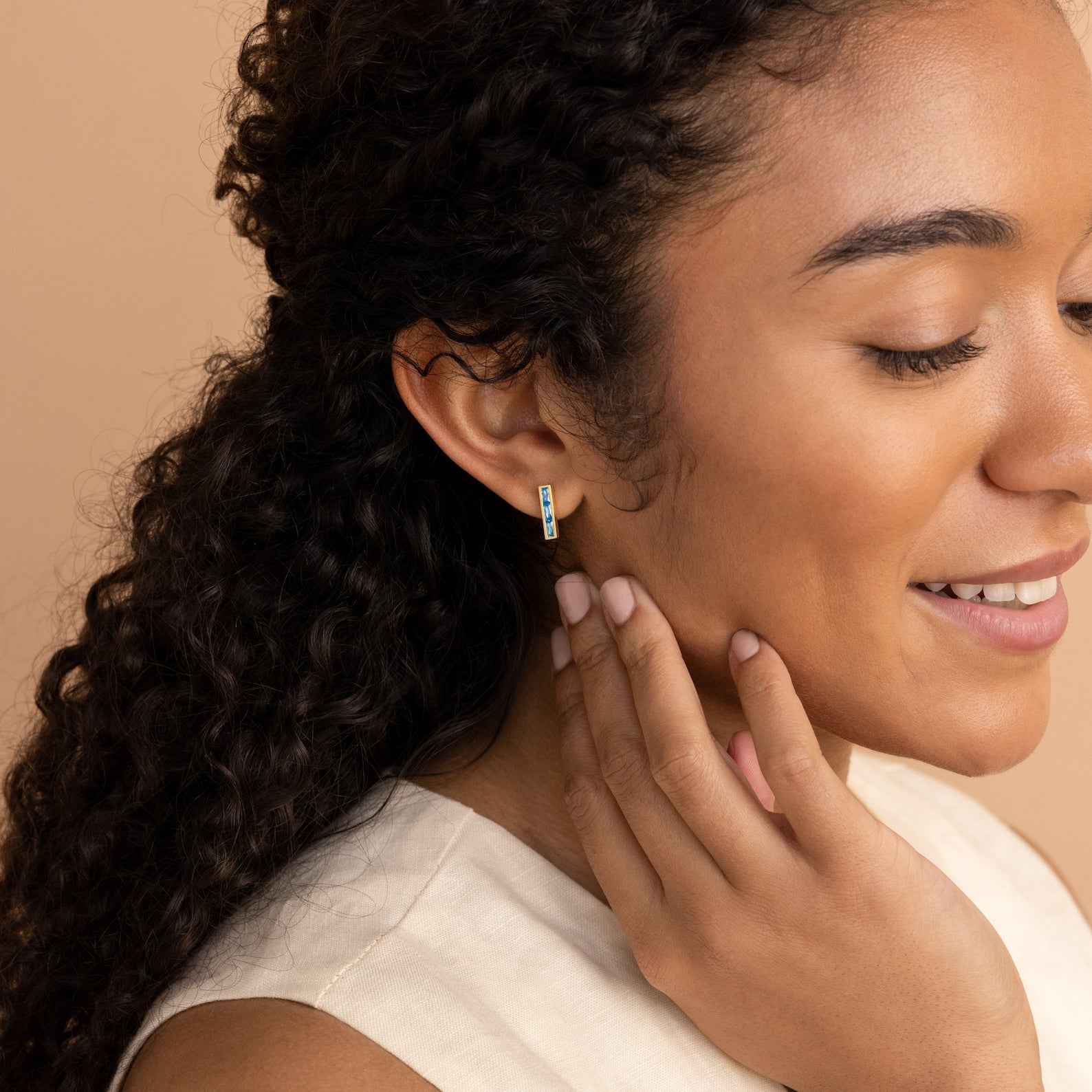 A smiling woman with curly hair touches her ear, displaying our Baguette Birthstone Bar Studs—a small gold hoop earring with gems and a delicate bar stud for her piercing.