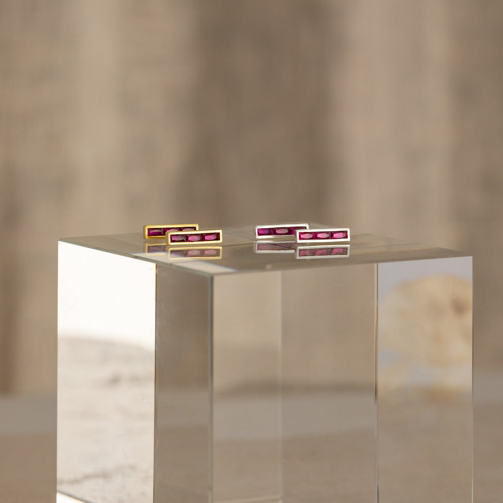 The Baguette Birthstone Bar Studs, featuring four rectangular bars with pink stones, are elegantly displayed on a clear block pedestal against a neutral background.