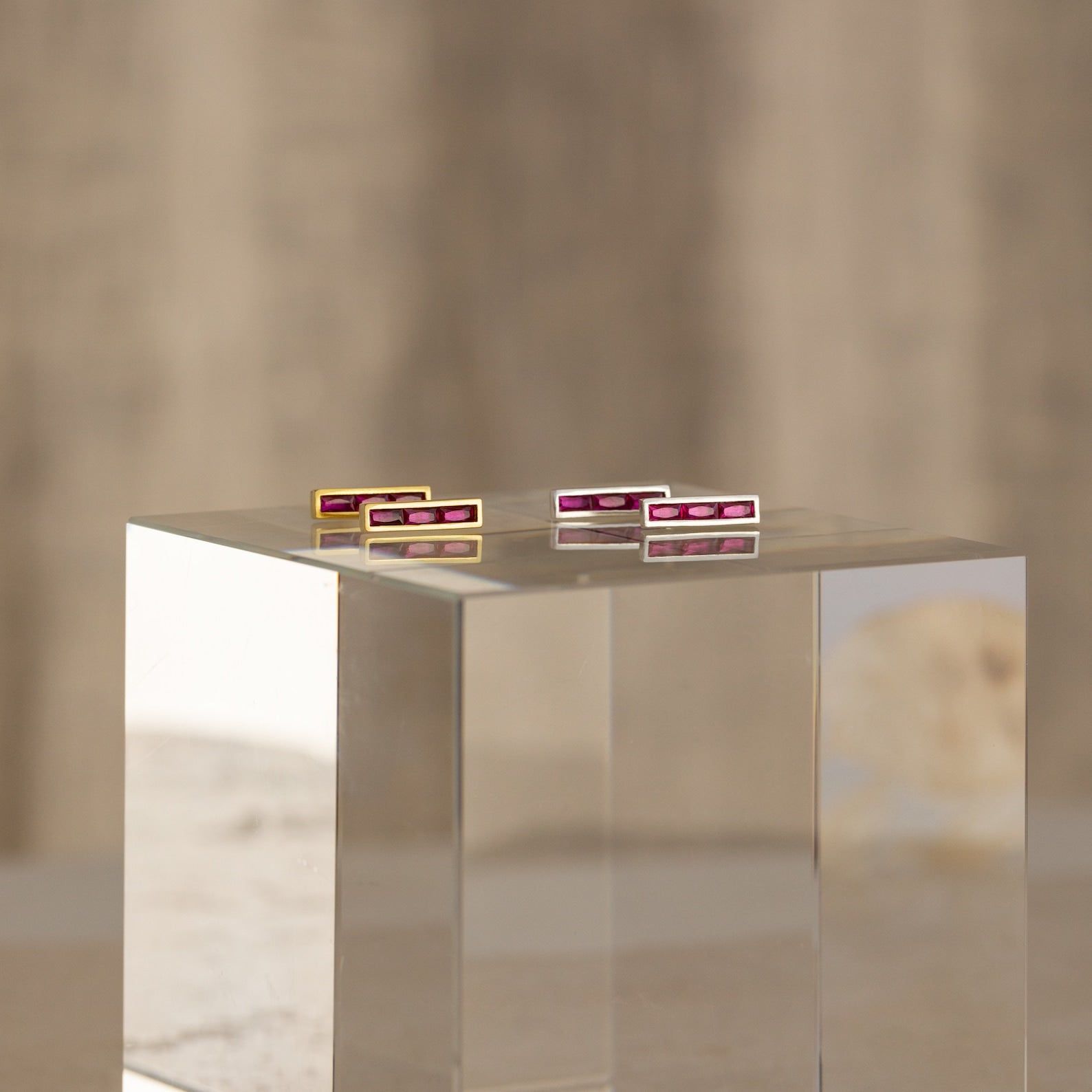 The Baguette Birthstone Bar Studs, featuring four rectangular bars with pink stones, are elegantly displayed on a clear block pedestal against a neutral background.