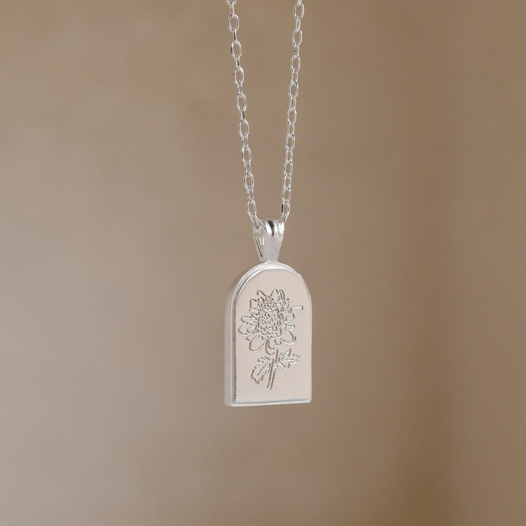 The Birth Flower Arch Necklace features an engraved floral design in silver, displayed against a neutral beige background—a stunning piece of personalized jewelry.